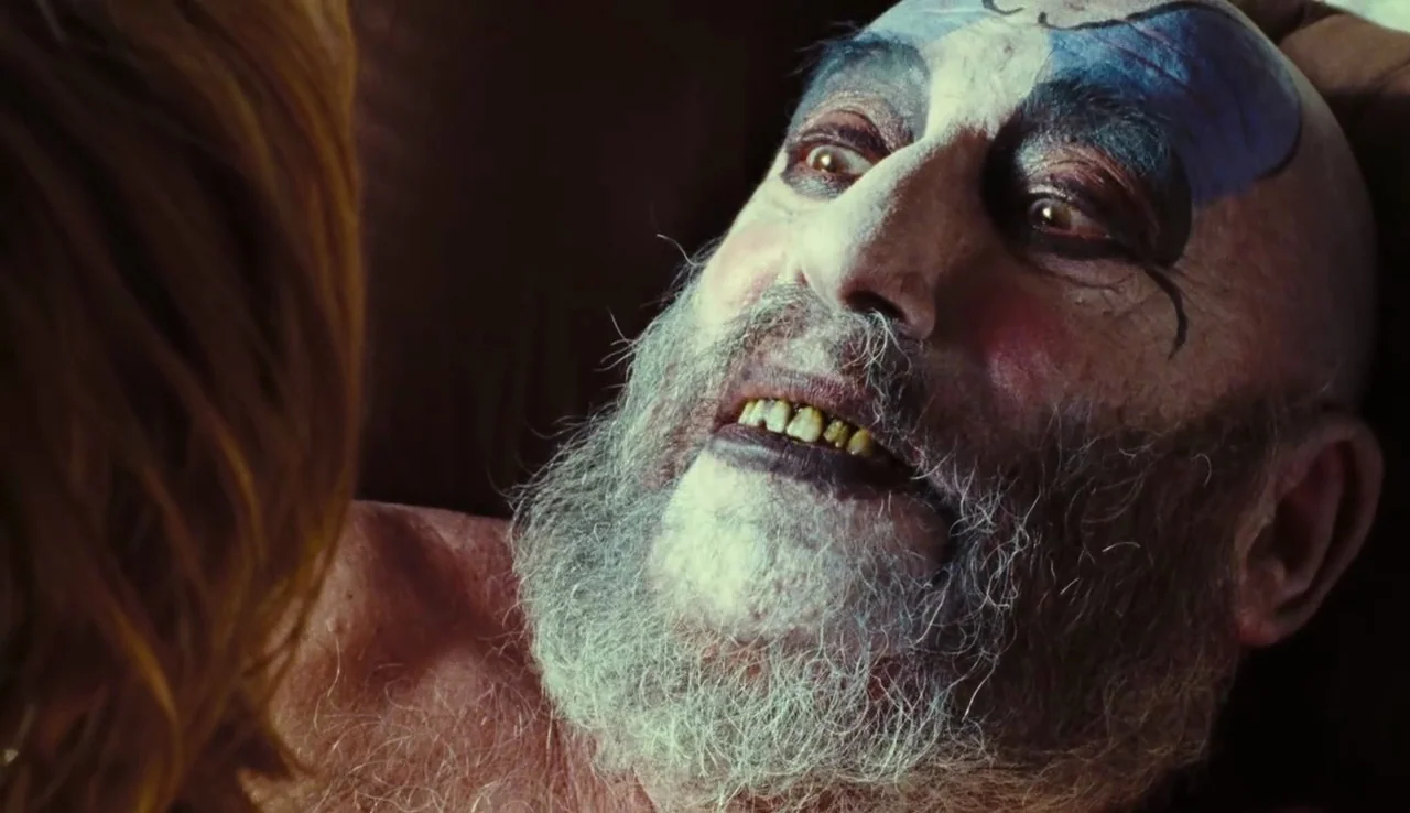 The Devil’s Rejects (2005) movie still: closeup — A dimly lit indoor space with shadows obscuring the background. An elderly man with white clown…