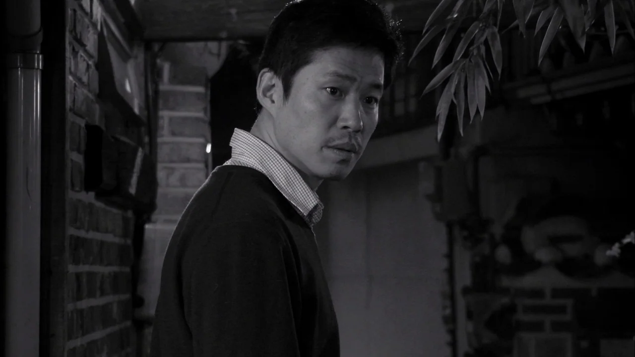 The Day He Arrives (2018) movie still: medium — A narrow residential passage features a brick wall on the left side. A man turns his head while…