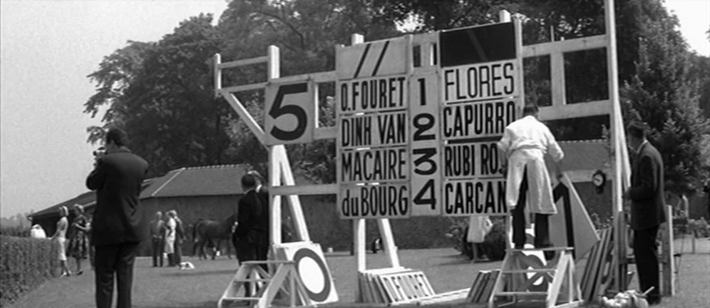 Les Amants (1958) movie still: wide — A grassy field at a racetrack features a large wooden board listing horse names. A man wearing a white coat…