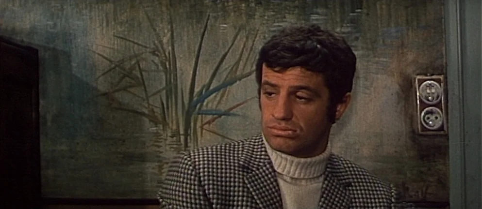Mississippi Mermaid (1969) movie still: medium — A man stands indoors before a wall painted with a mural of tall reeds. He gazes off-camera while…