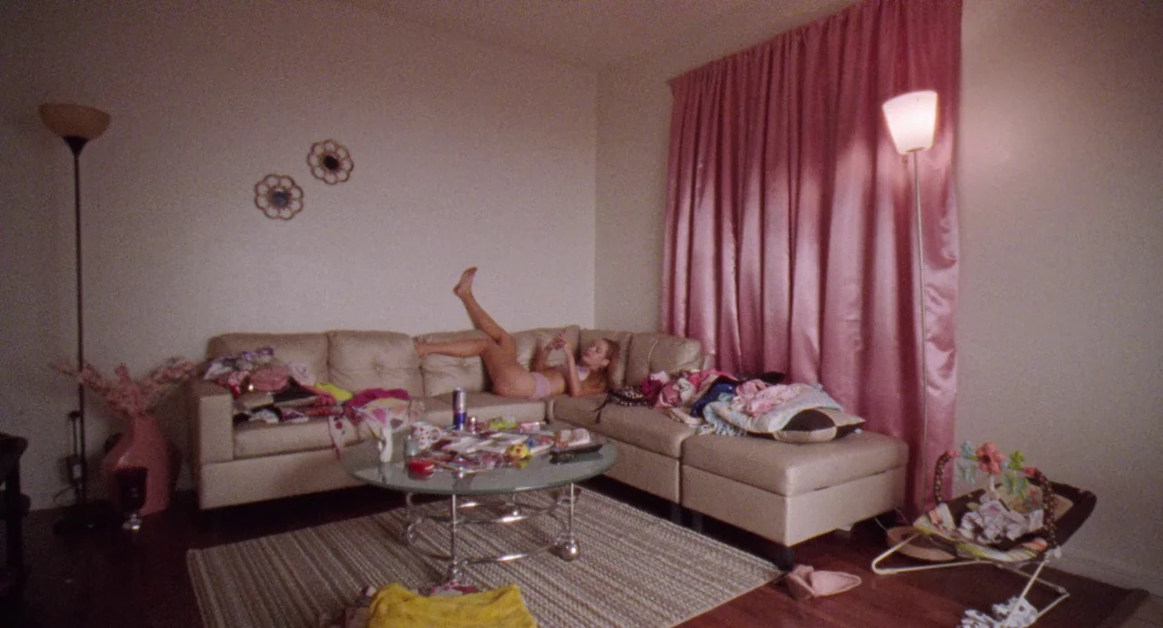 Zola (2020) movie still: wide — A cluttered apartment space features a large beige sectional sofa and a glass coffee table. A young woman lies on…
