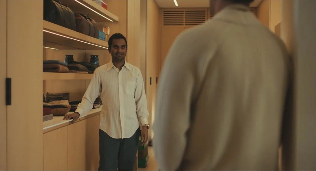 Good Fortune (2025) movie still: medium — A well-lit, organized walk-in closet features light-wood shelving and neatly stored items. A man in a…