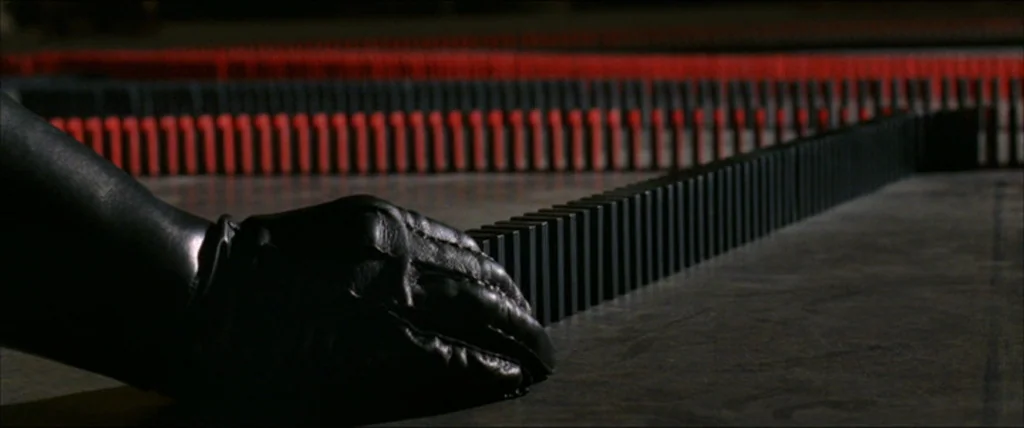 V for Vendetta (2006) movie still: closeup — A dark concrete floor features a long, winding row of black and red dominoes. A hand wearing a black…