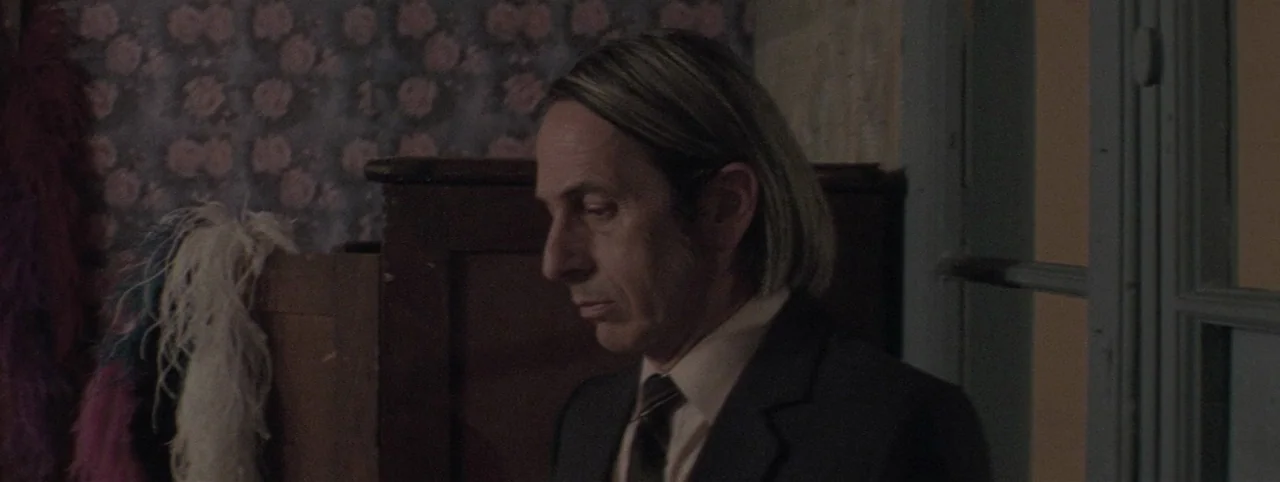 Post Mortem (2010) movie still: medium — A dimly lit room features patterned wallpaper with floral motifs and hanging colorful feathers. A man in a…
