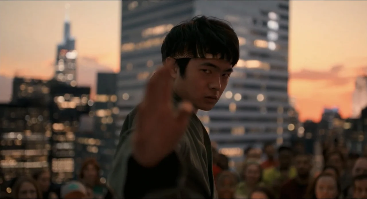 Karate Kid: Legends (2025) movie still: medium — A rooftop terrace overlooking a dense city skyline at sunset. A young East Asian man in a martial…