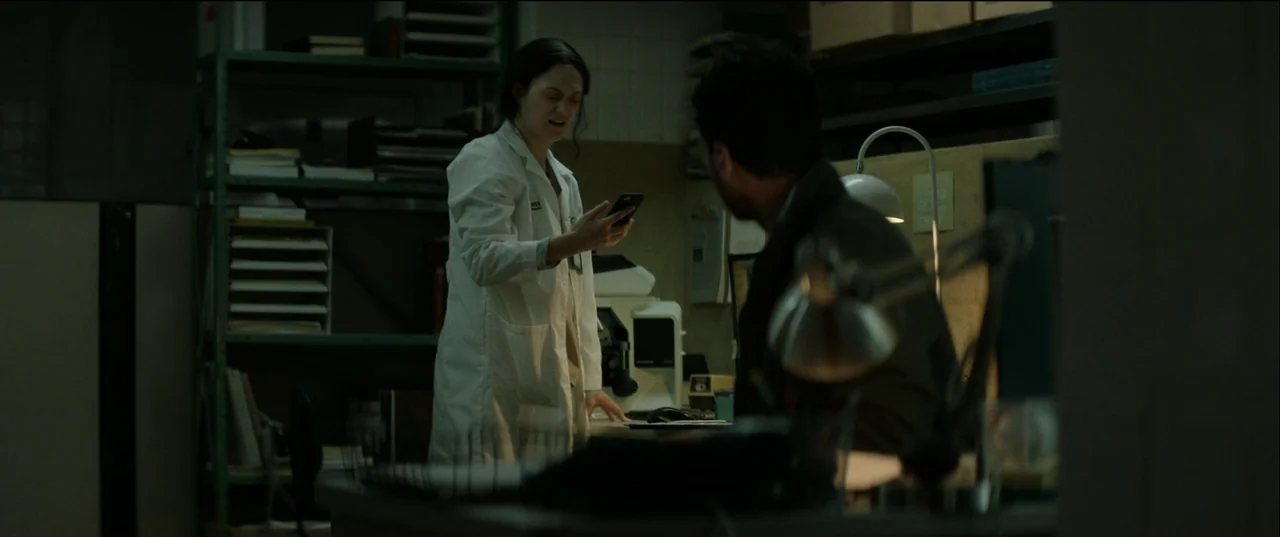 Birth/Rebirth (2023) movie still: medium — A dimly lit room with shelves containing files and laboratory equipment in the background. A female…
