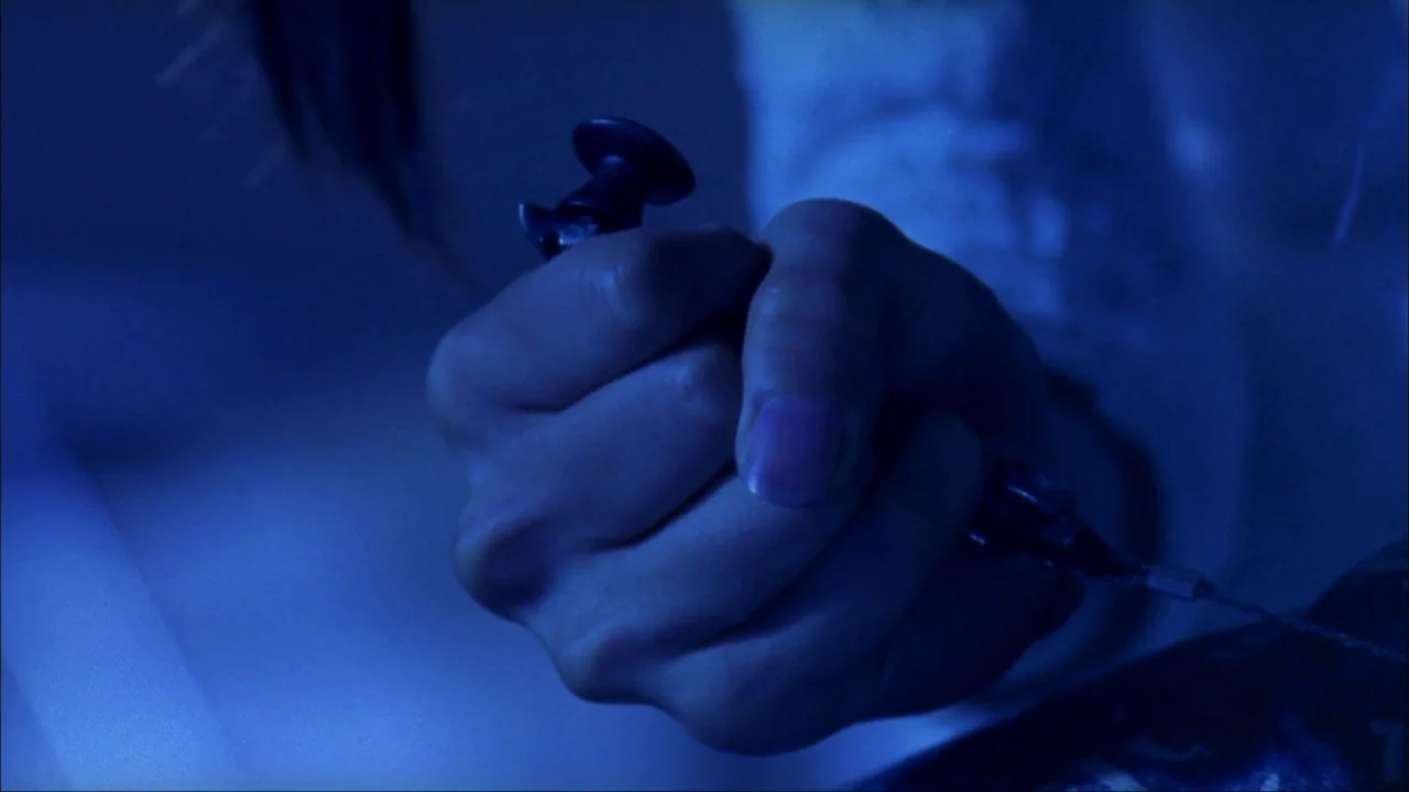 Audition (1999) movie still: closeup — A close-up captures a clenched hand against a dark, blue-lit background. The fingers grasp a black plastic…