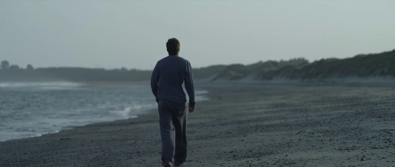 What Richard Did (2012) movie still: fullbody — A wide sandy beach stretches along the coast under a hazy, overcast sky. A man walks away from the…