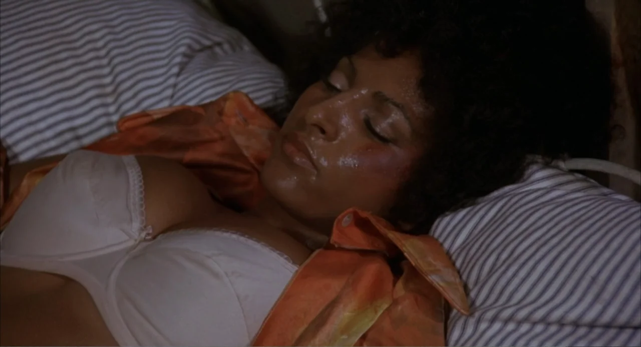 Foxy Brown (1974) movie still: closeup — A dimly lit bedroom features striped fabric pillows and a dark headboard. A woman with dark curly hair…
