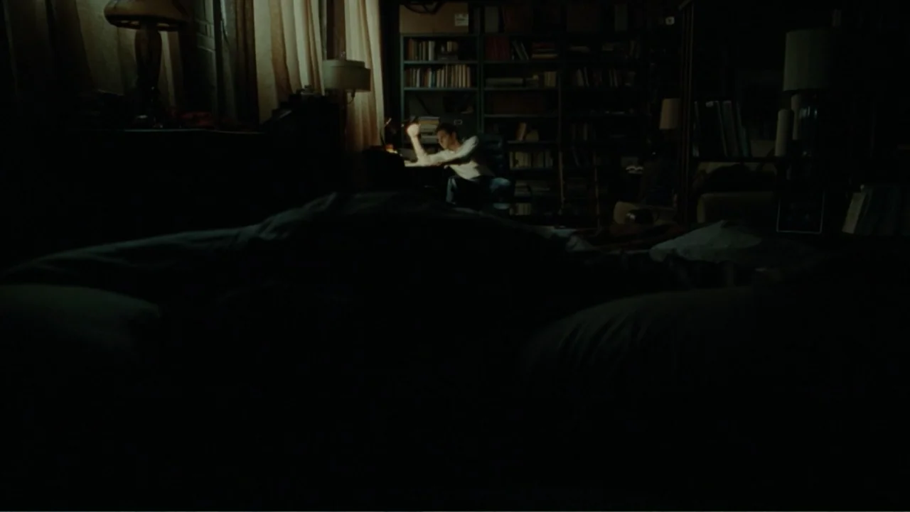 The Words (2012) movie still: wide — A dimly lit room filled with floor-to-ceiling bookshelves and heavy shadows. A man sits at a desk in the…