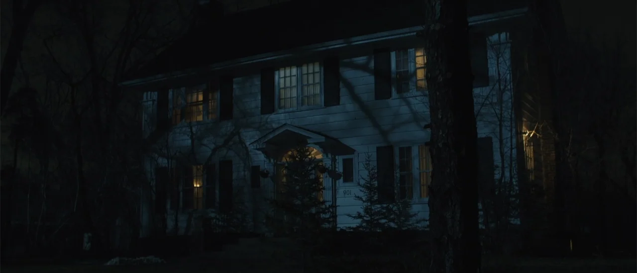 All Fun and Games (2023) movie still: wide — A two-story white suburban house at night with dark shutters and bare tree silhouettes. The windows…