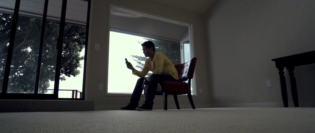 I Melt With You (2011) movie still: fullbody — A sparsely furnished room features carpeted floors and a large window looking outside. A man sits in…