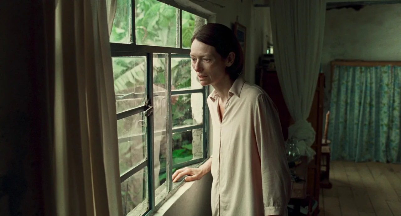 Memoria (2021) movie still: medium — A modest interior room features a white-framed window looking out to green foliage. A woman stands by the…