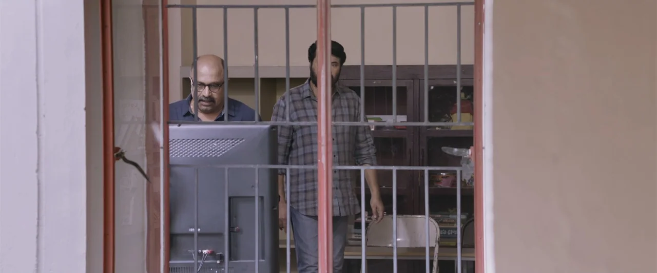 Peranbu (2019) movie still: medium — A utilitarian room features a bookshelf, a metal folding chair, and a flat-screen television. Two men stand…
