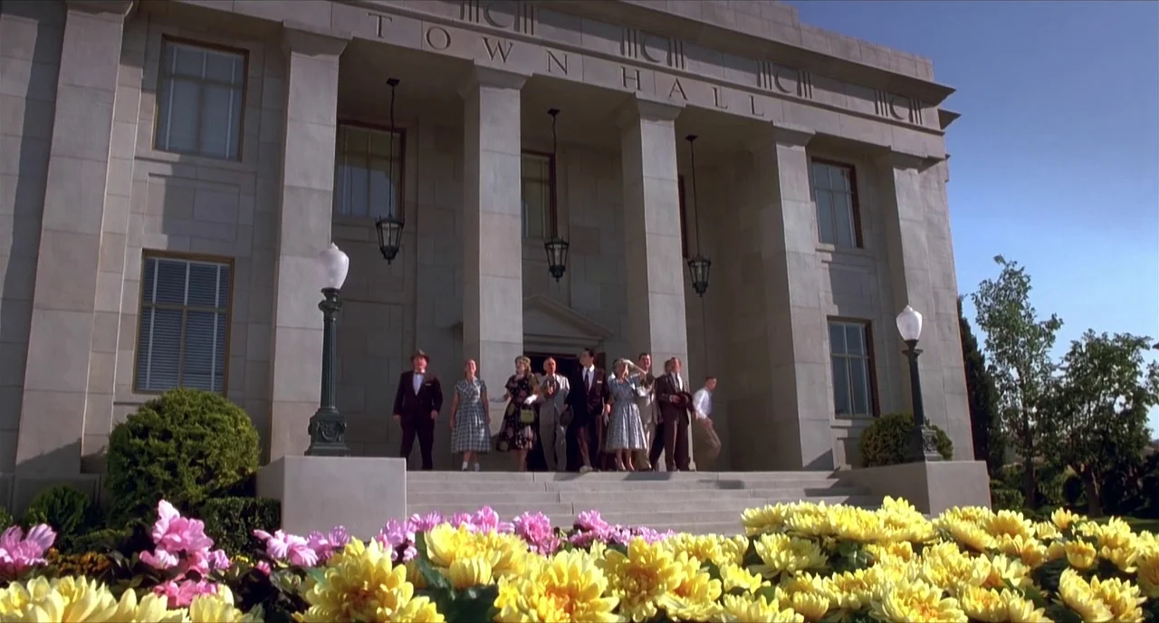 Pleasantville (1998) movie still: wide — A stone town hall with classic columns serves as a backdrop to a group. People in mid-century attire emerge…