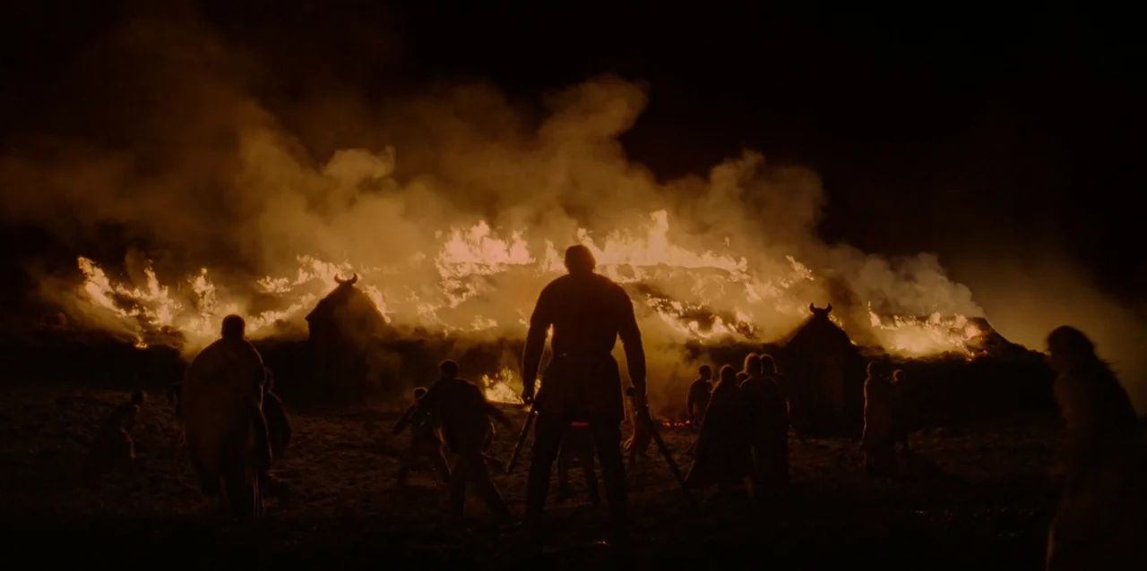 The Northman (2022) movie still: wide — A dark night landscape features several wooden longhouses with horned roofs engulfed in flames. A group of…