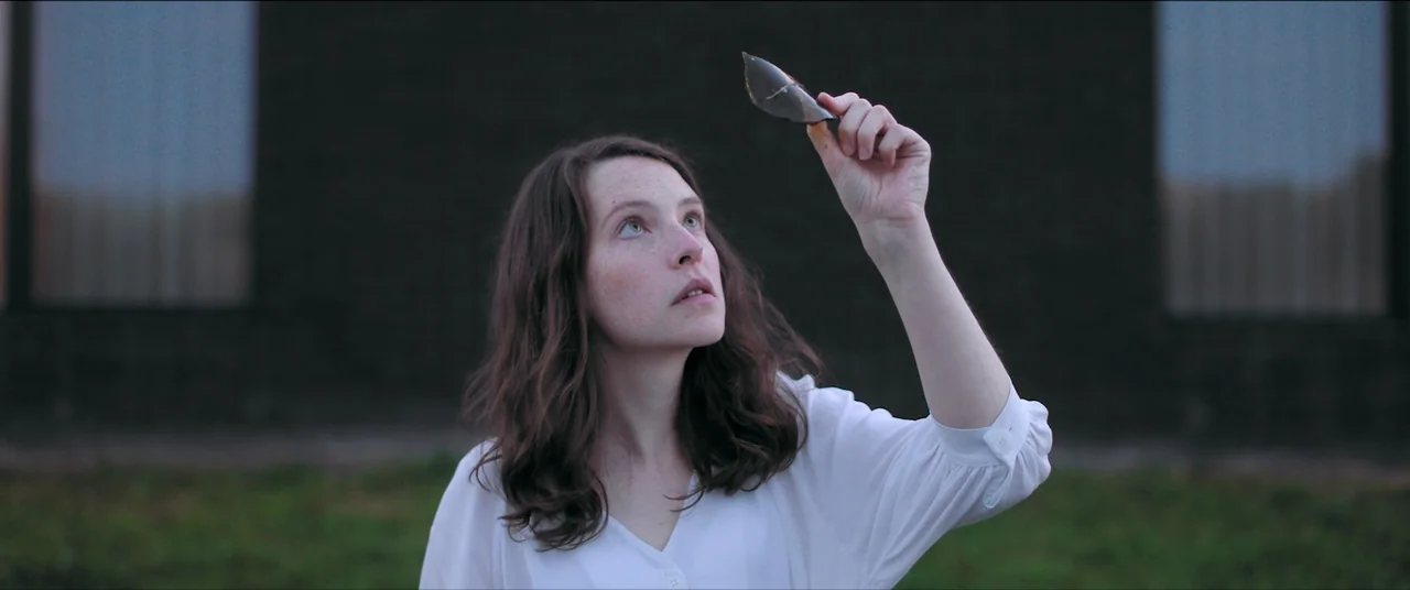 The Feast (2021) movie still: medium — A woman stands in front of a dark, plain building exterior during blue hour. She holds a small, sharp shard…