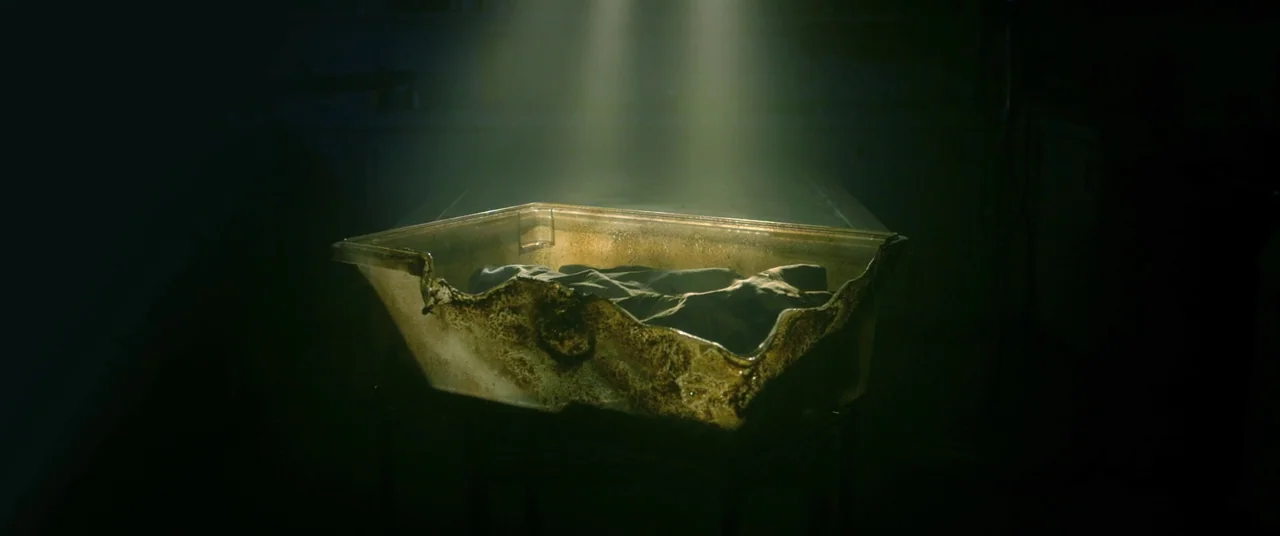 The Void (2016) movie still: medium — A translucent plastic container rests on a metal stand within a dark room. The container is stained with dark…