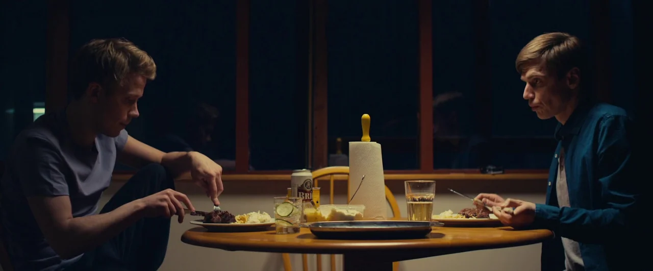Rift (2017) movie still: medium — Two men sit across from each other at a wooden table in a dark room. They are eating a meal with plates, glasses,…