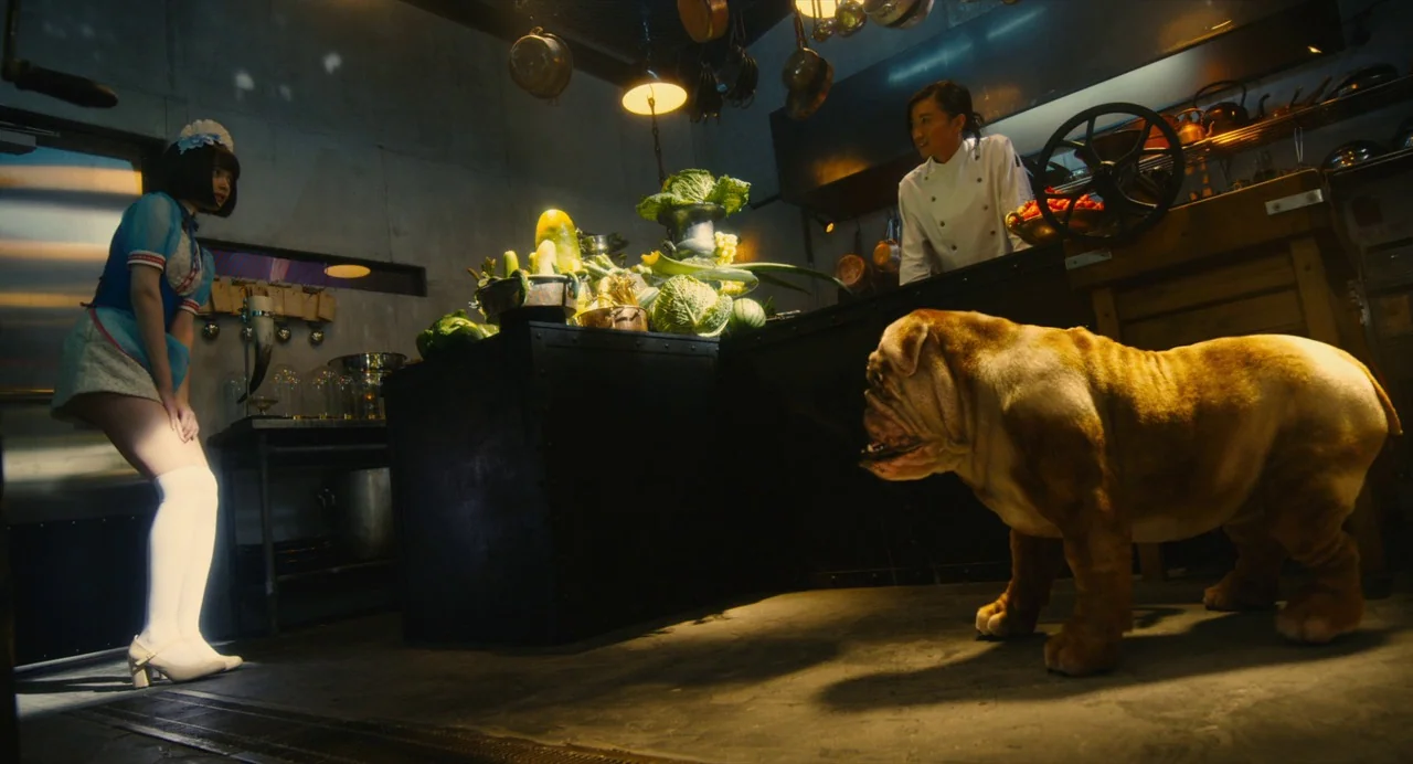 Diner (2019) movie still: fullbody — A modern industrial kitchen with concrete walls and hanging copper pans over a dark counter. A young woman in a…
