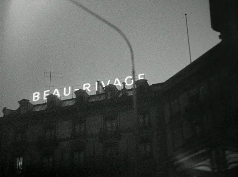 Le Petit Soldat (1963) movie still: wide — A multistory building with a glowing rooftop sign stands against a dark evening sky. The neon sign…