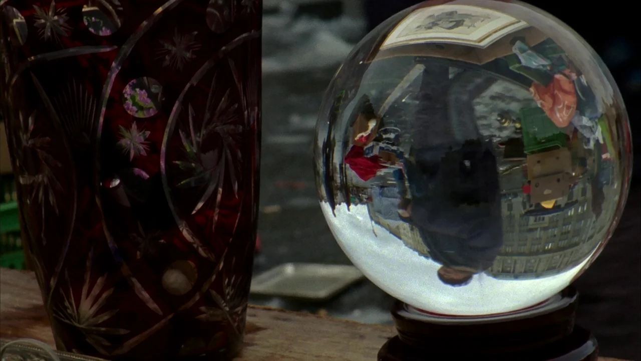 Museum Hours (2012) movie still: closeup — A cut glass red vase stands next to a clear crystal sphere on a table. The crystal sphere shows an…