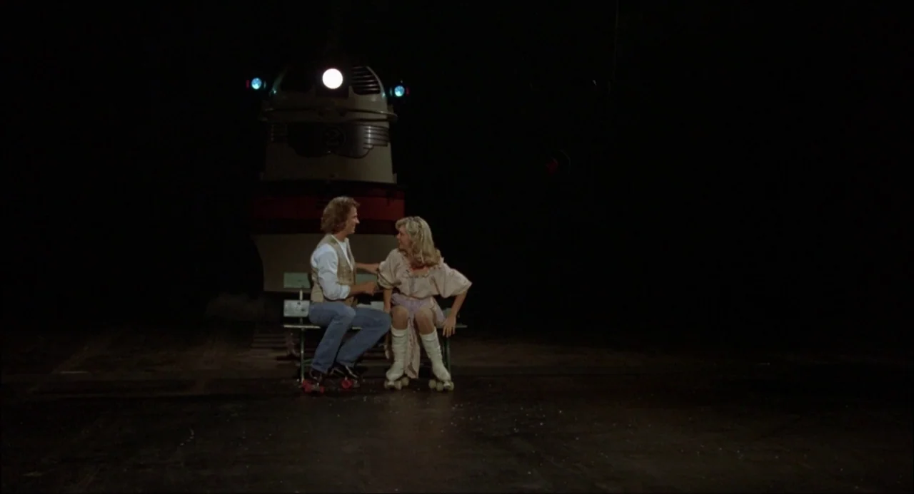Xanadu (1980) movie still: fullbody — A man and woman sit on a metal bench in an expansive black void. A large, stylized train-like vehicle with…