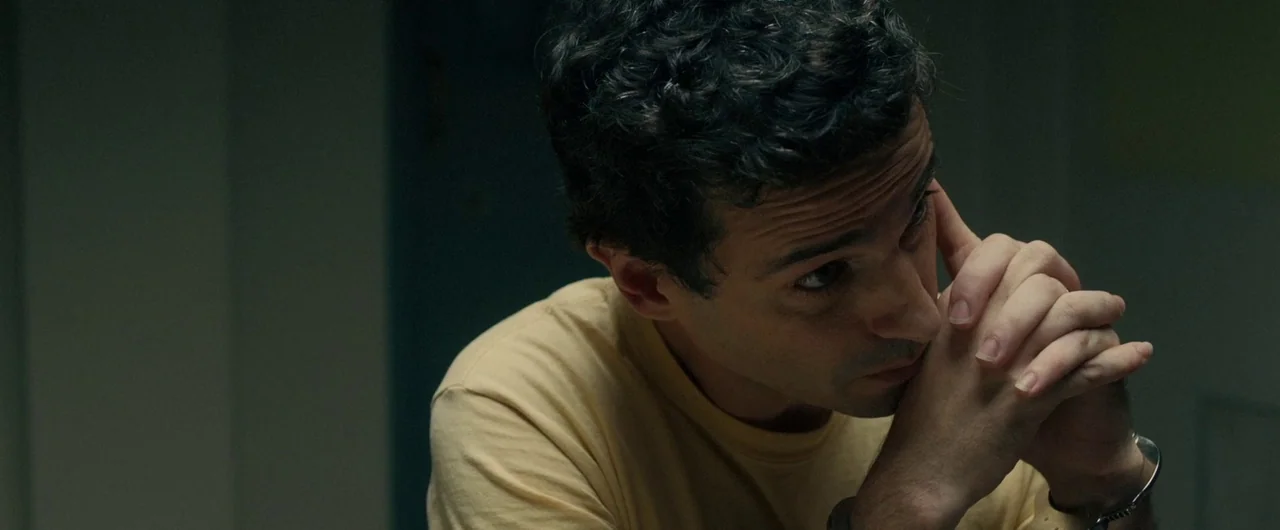 No Man of God (2021) movie still: closeup — A dimly lit room with dark walls and minimal environmental detail. A man in a yellow t-shirt sits with…