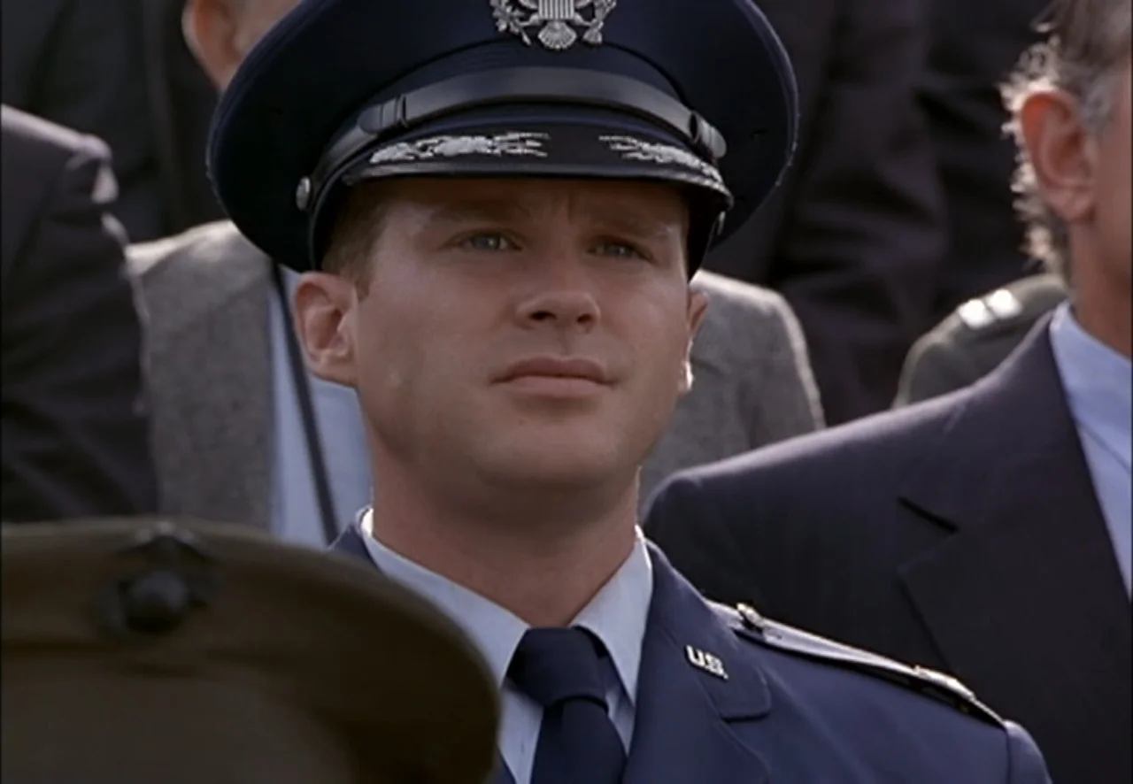 The Pentagon Wars (1998) movie still: medium — A group of people wearing formal attire are assembled in an open grassy area. An Air Force officer in…