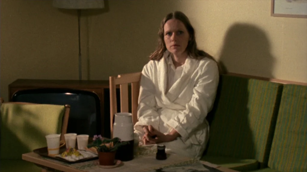 Face to Face (1976) movie still: medium — A woman in a white bathrobe sits on a green sofa in a room. A small table with a tray and a drink stands…