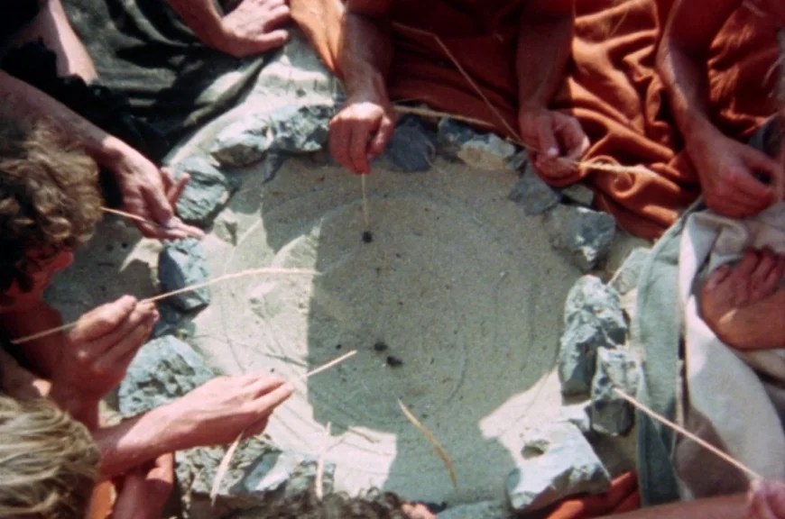 Sebastiane (1976) movie still: medium — A circle of grey rocks is arranged on a patch of loose sand. Several people sit around the circle using…