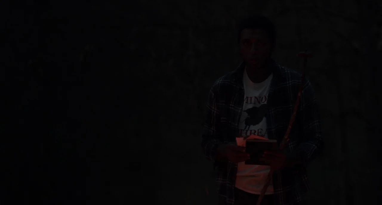 The Alchemist Cookbook (2016) movie still: medium — A dark outdoor setting at night obscures the surroundings with heavy shadows. A young man stands…