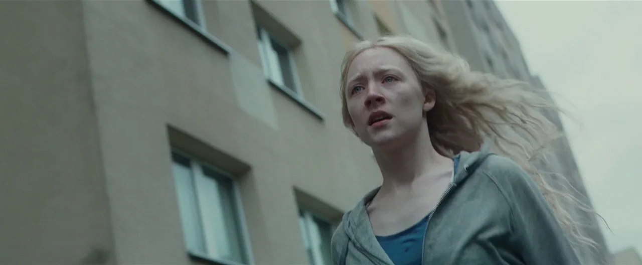 Hanna (2011) movie still: medium — A woman stands before the exterior of a grey concrete apartment building. She looks forward while her long hair…