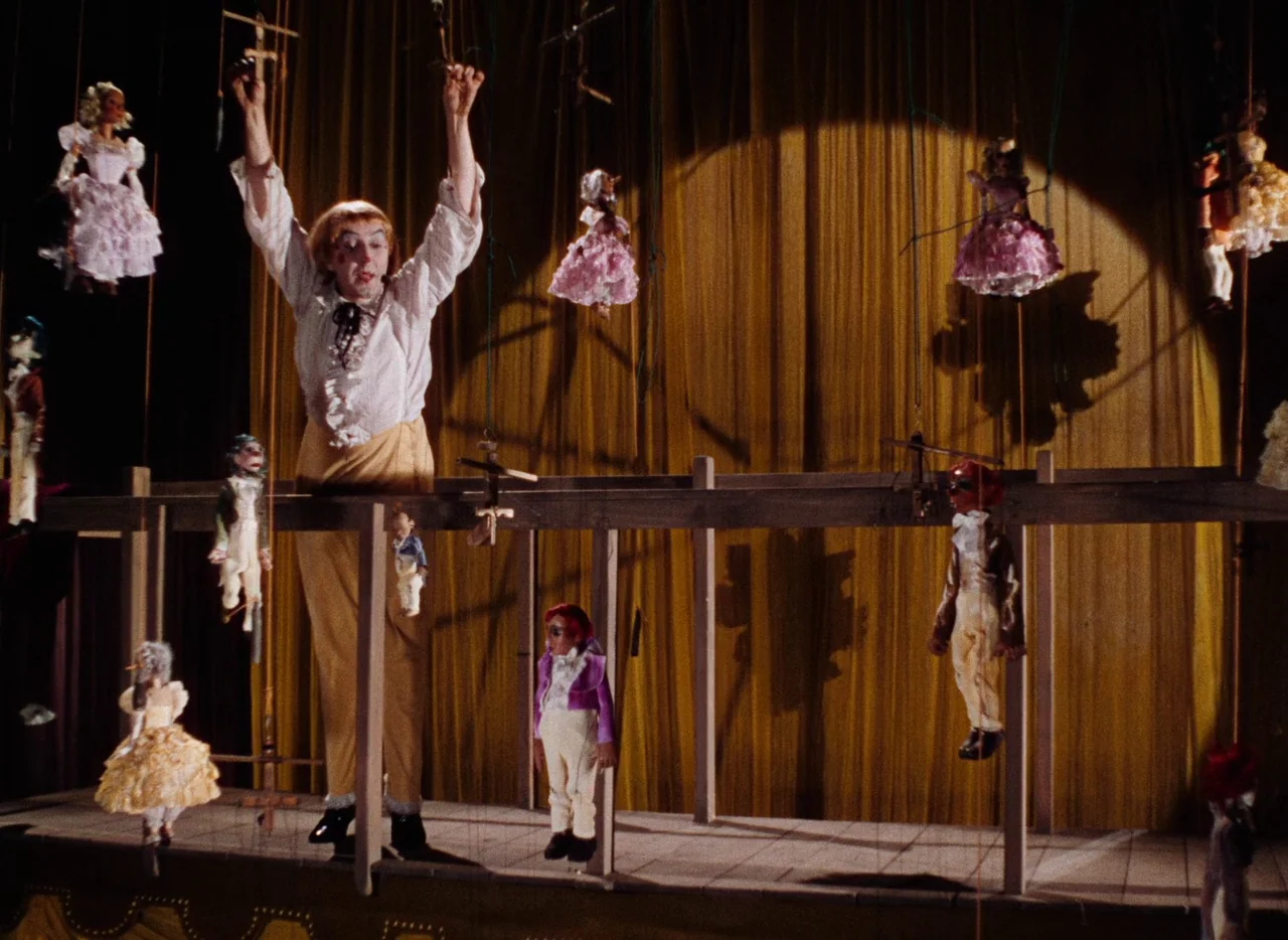 The Tales of Hoffmann (1951) movie still: fullbody — A wooden platform with multiple hanging marionettes sits against a draped golden-yellow velvet…