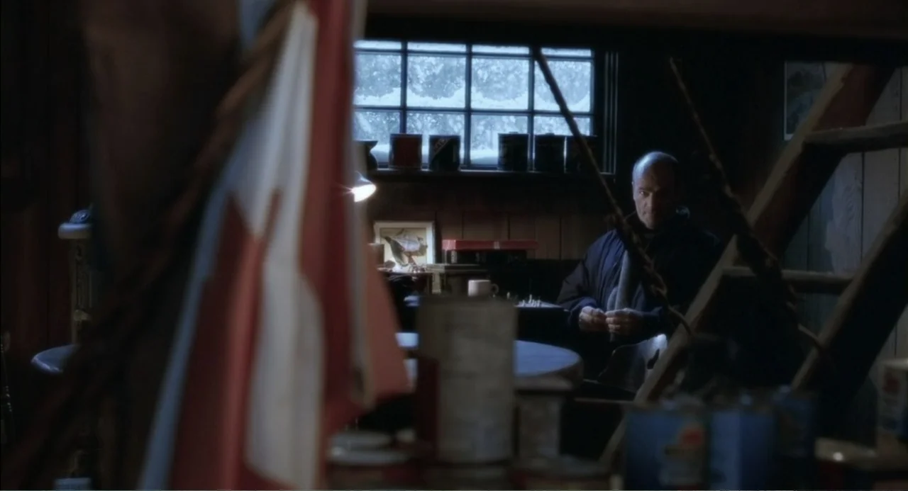 The Hunted (2003) movie still: medium — A dimly lit cabin interior features wooden walls and a window overlooking snow. A man sits at a small table…