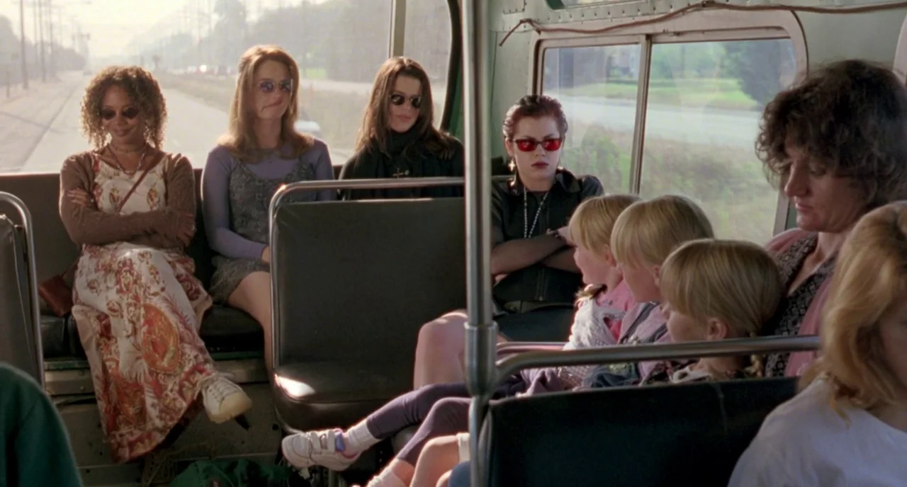 The Craft (1996) movie still: medium — A group of young women sit together in the back of a city bus. They look forward while children sit in the…