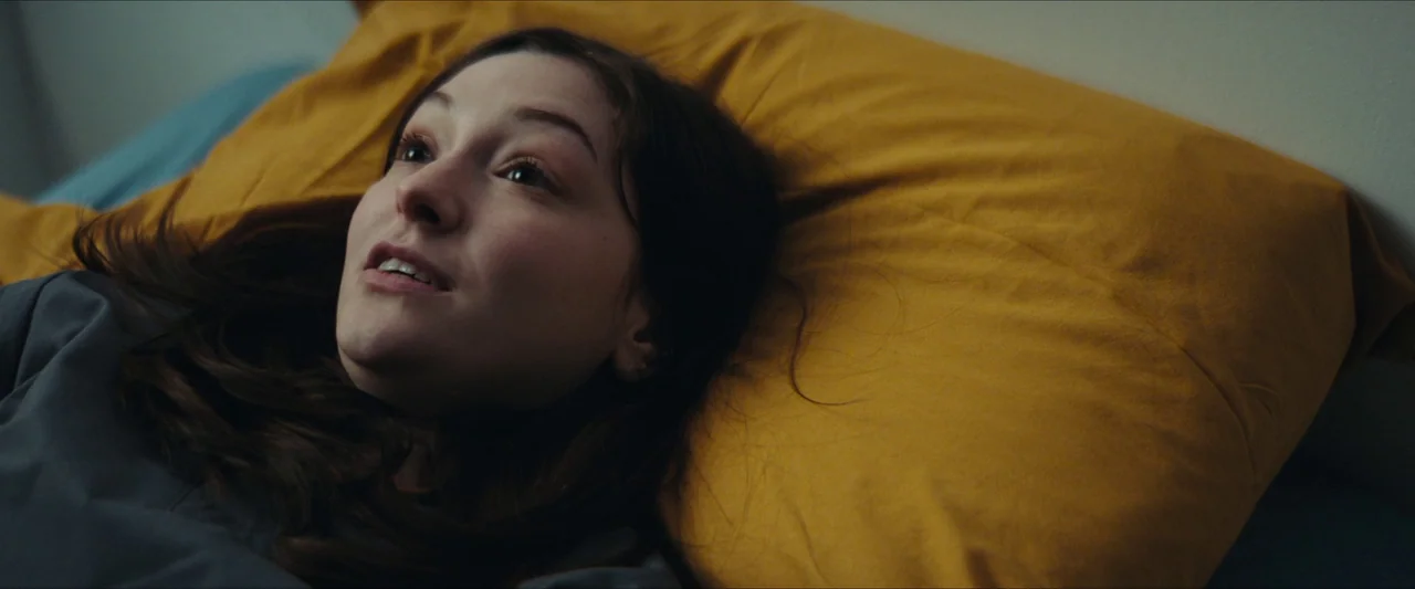 Double Blind (2023) movie still: closeup — A person lies on a vibrant yellow pillow on a bed. The individual looks upward toward the side with an…