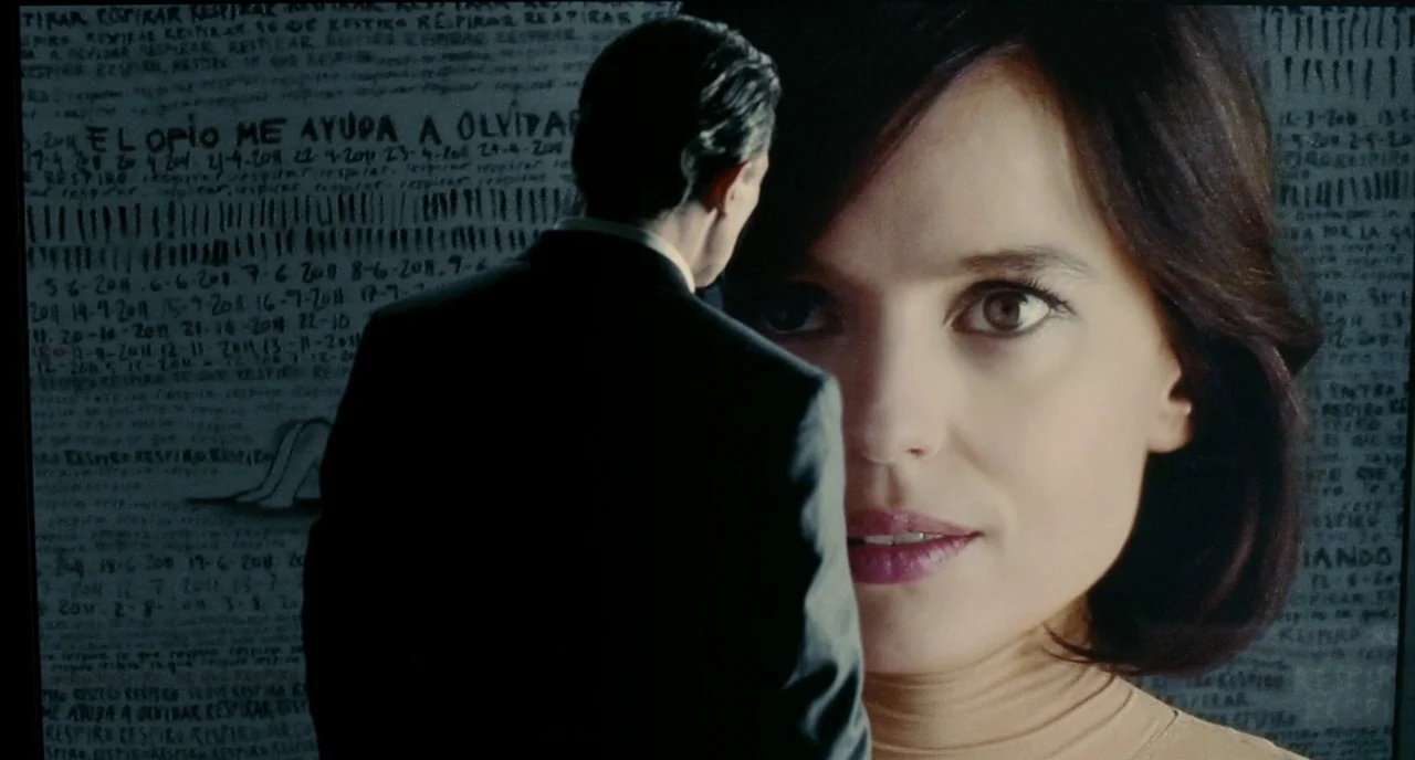 The Skin I Live In (2011) movie still: medium — A man stands before a dark wall covered in repeated handwritten lists and dates. A large-scale video…