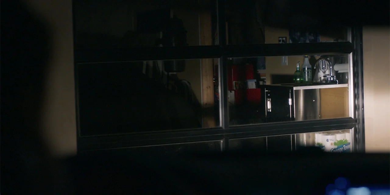 Demonic (2015) movie still: wide — A dimly lit garage interior is viewed through a glass-paneled partition. The visible area contains a metal…