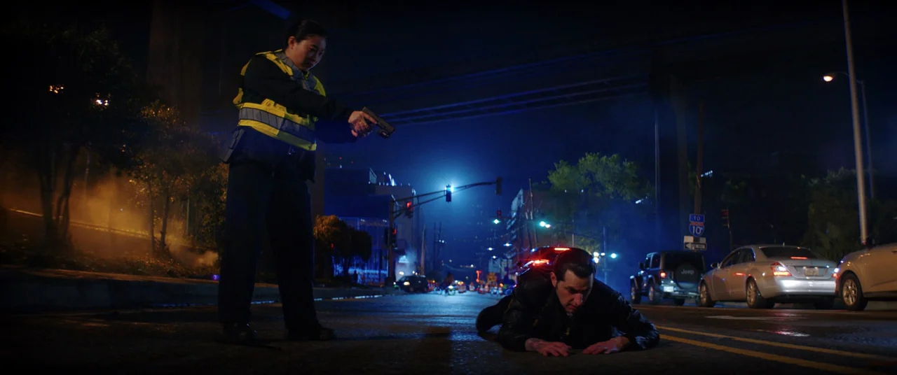 Renfield (2023) movie still: wide — A night scene on an asphalt road located under a concrete highway overpass. A police officer stands aiming a…