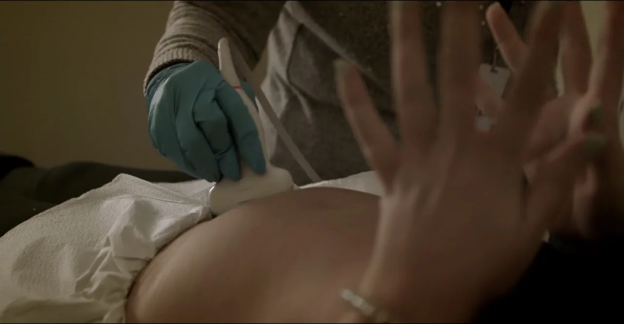 The Devil Is Busy (2024) movie still: closeup — A clinical examination room with white fabric covering a patient's abdomen. A medical professional…