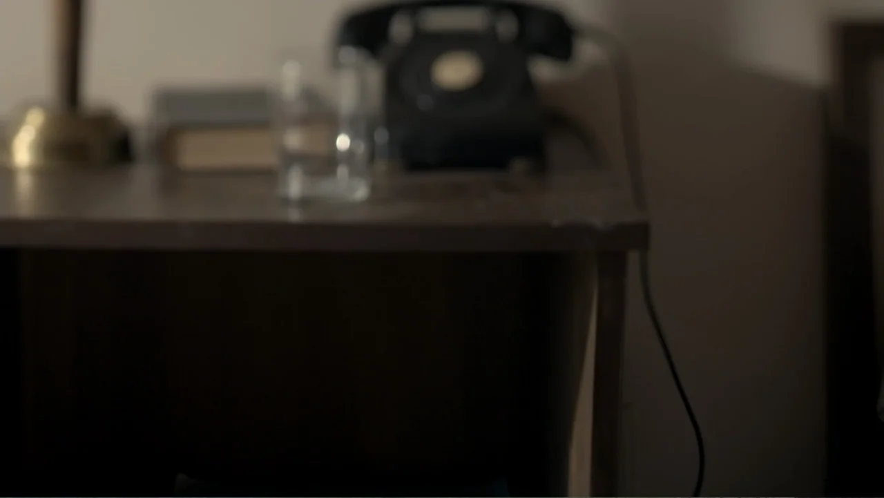 King in the Wilderness (2018) movie still: closeup — A dark wooden side table holds a vintage telephone and a glass. No occupants are present in…