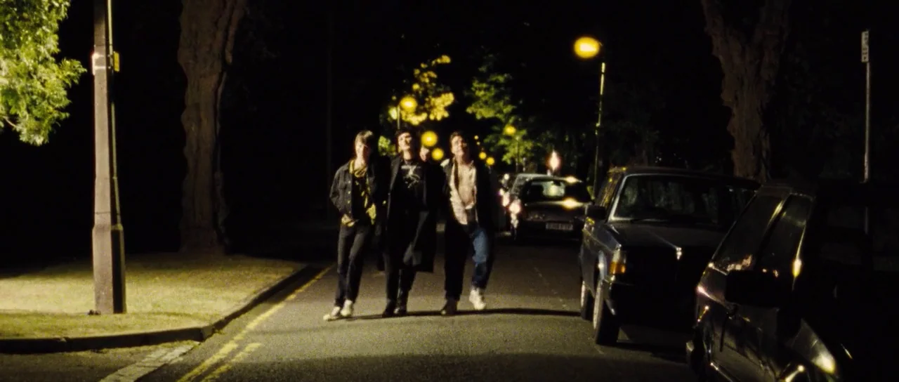 The World’s End (2013) movie still: fullbody — Three young men walk arm-in-arm down the middle of a quiet residential street at night. Cars are…