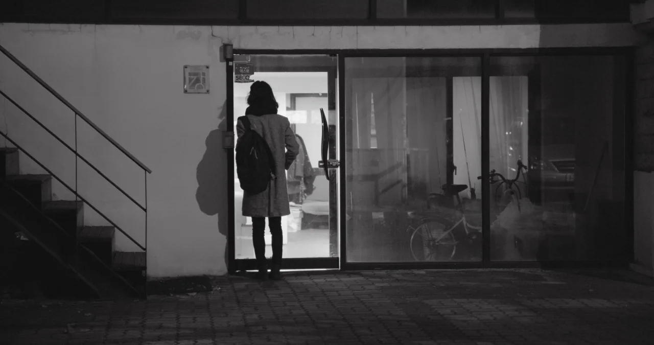 The Day After movie still: fullbody — A building exterior at night features a glass door illuminated from within the interior. A person with a…