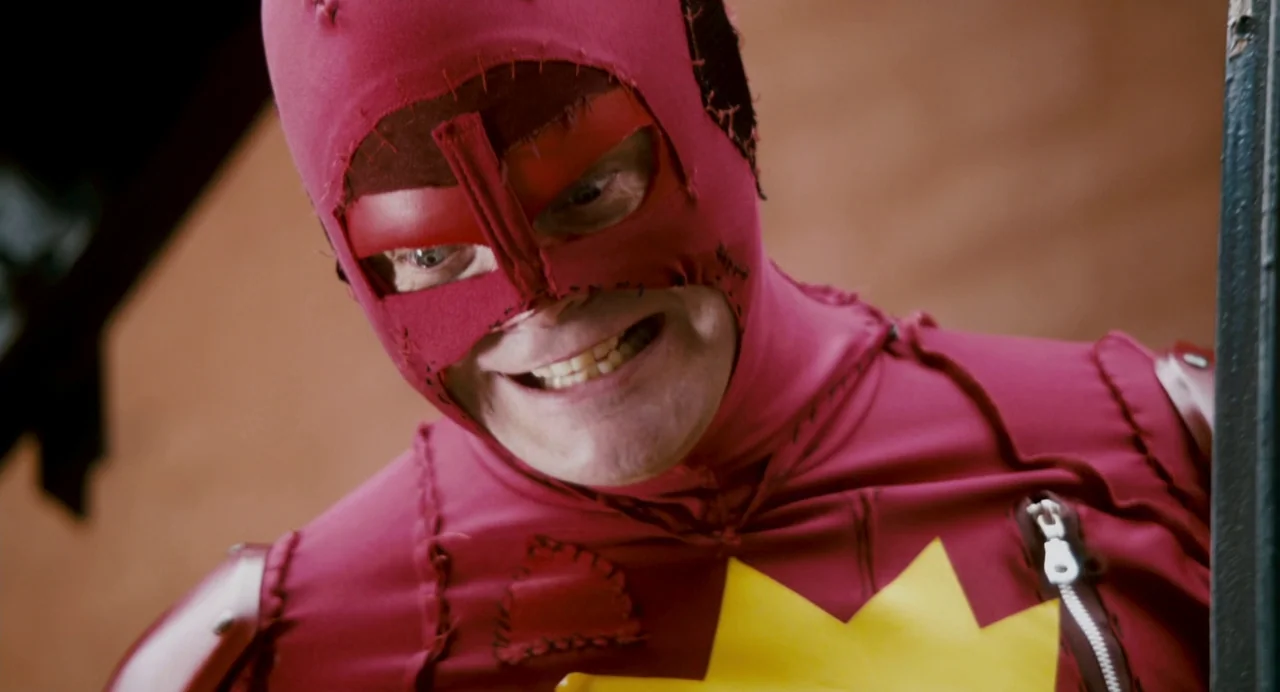 Super (2010) movie still: closeup — A man wears a DIY red fabric costume with stitched panels and a star emblem. He smiles directly at the viewer…