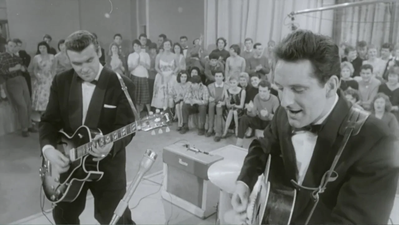Becoming Led Zeppelin (2025) movie still: medium — A television studio features two guitarists in tuxedos performing for a seated audience. The band…