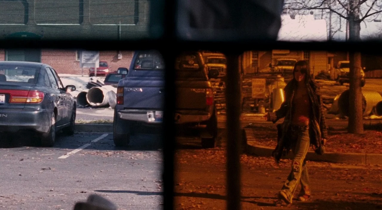 Halloween 2 (2009) movie still: wide — A paved lot sits between buildings with several parked vehicles and sparse trees. A young woman walks…