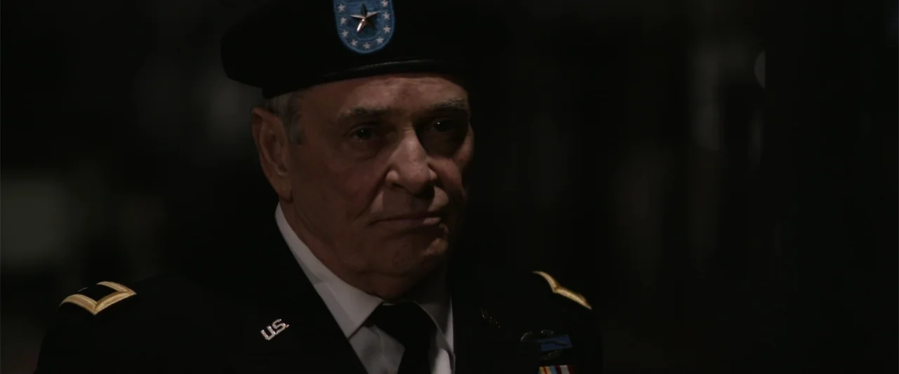 The Condemned 2 (2015) movie still: closeup — A man in a military dress uniform stands in a dark, indistinct setting. The subject stares off-screen…
