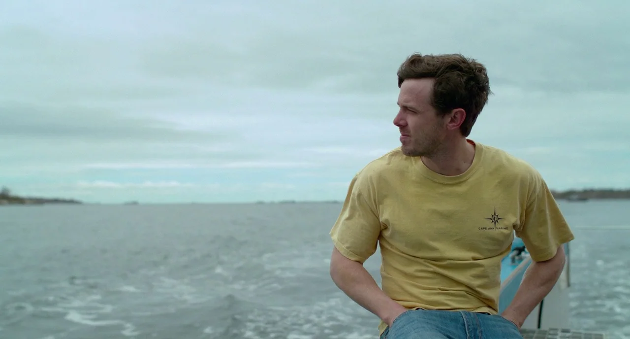 Manchester By The Sea (2016) movie still: medium — A man sits on a boat deck surrounded by vast open water under overcast skies. The man gazes…