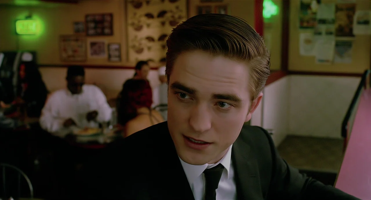 Cosmopolis (2012) movie still: closeup — A man in a suit sits in a dimly lit diner at night. He looks off-camera while other patrons are seated at…